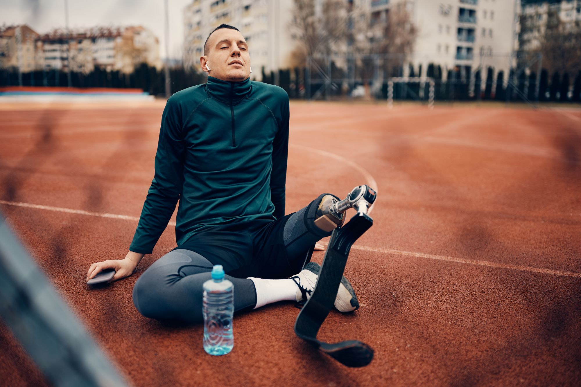 Young athletic man with artificial leg taking a break and listening music over wireless earphones at the stadium.