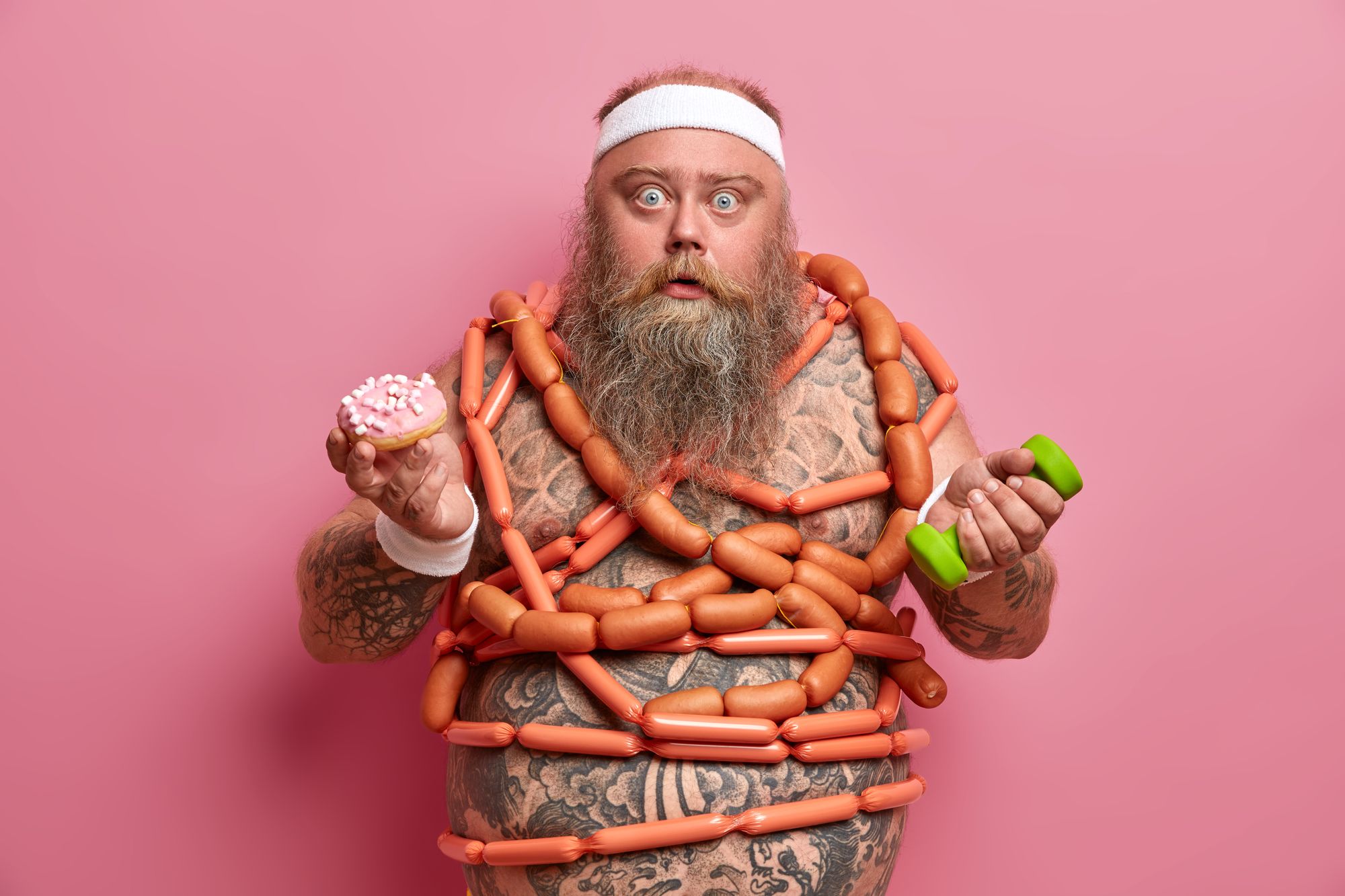 Stupefied stout bearded man poses with junk food and dumbbell, holds donut, many sausages around naked tattooed body