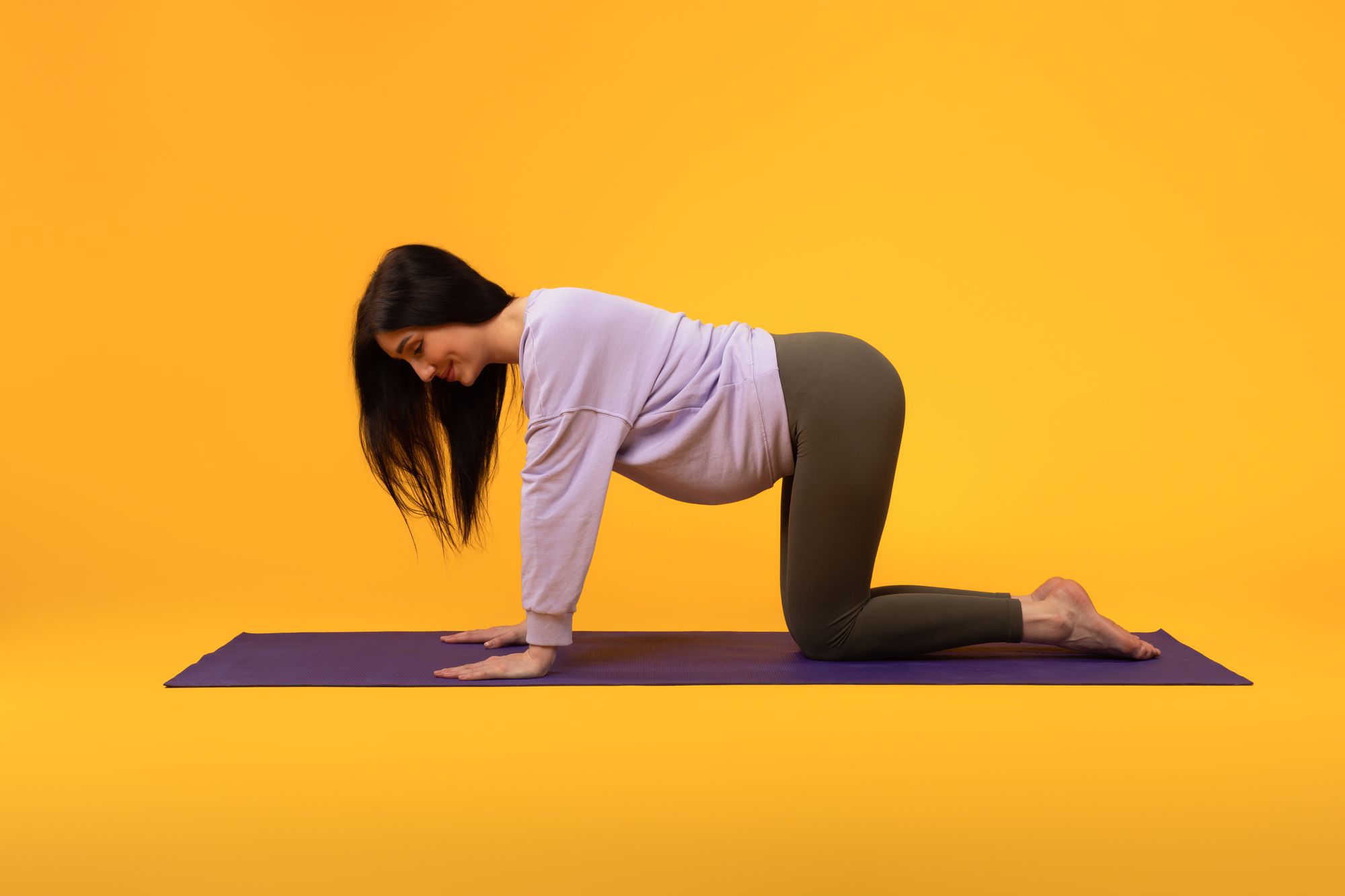 e view of young pregnant lady exercising on yoga mat, expectant woman in activewear practicing sports on yellow background