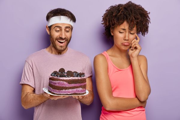 Upset woman turns from husband who holds tasty cake on plate, has sad expression as cannot eat sweets