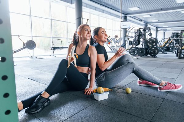 Two female friends in sportive clothes is in the gym earing fruits and taking a break