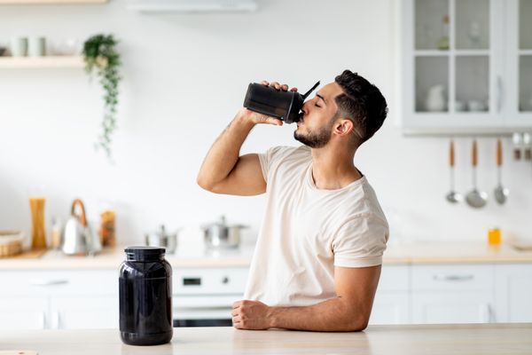 Young Arab guy drinking protein shake from bottle at kitchen, copy space